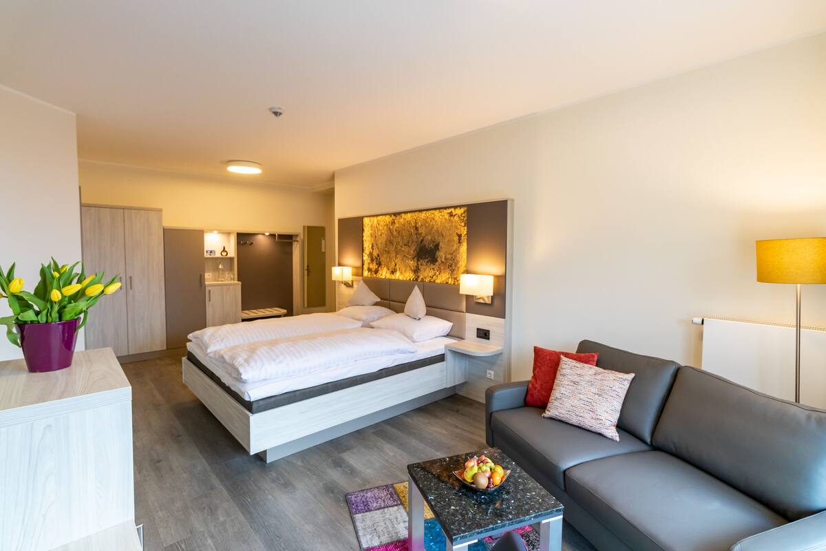 A modern hotel room with a bed, a gray sofa, a small coffee table with fruit, a potted plant, and a large abstract painting on the wall.