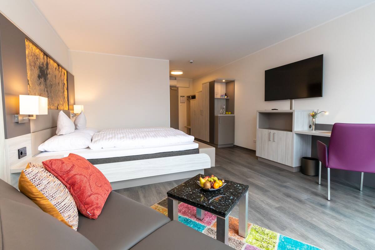 A modern hotel room with a bed, a small sofa, a desk, a TV, and a kitchen area.