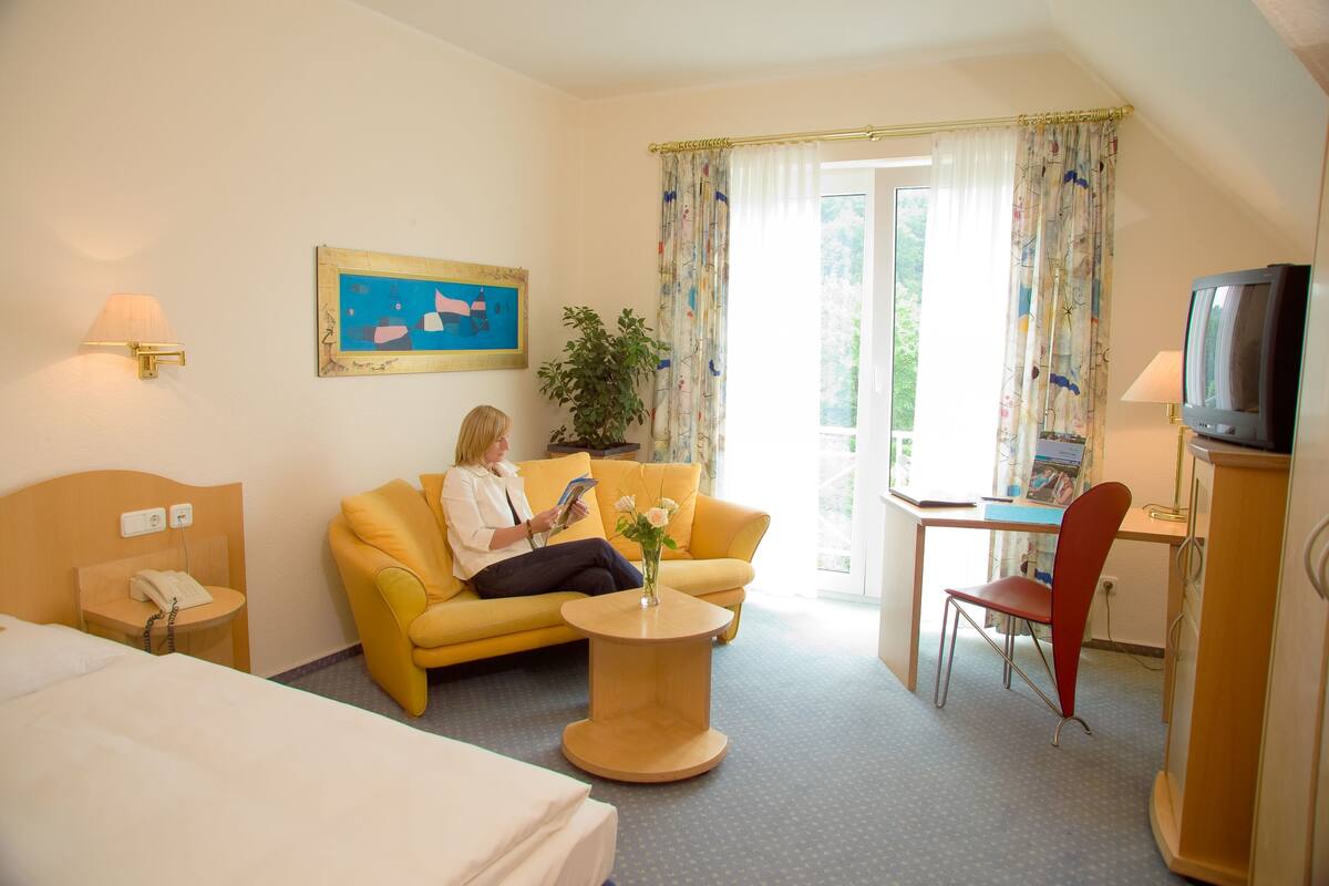 A hotel room with a bed, a yellow sofa, a small round table with a vase, a desk with a chair, a television, and a painting on the wall.