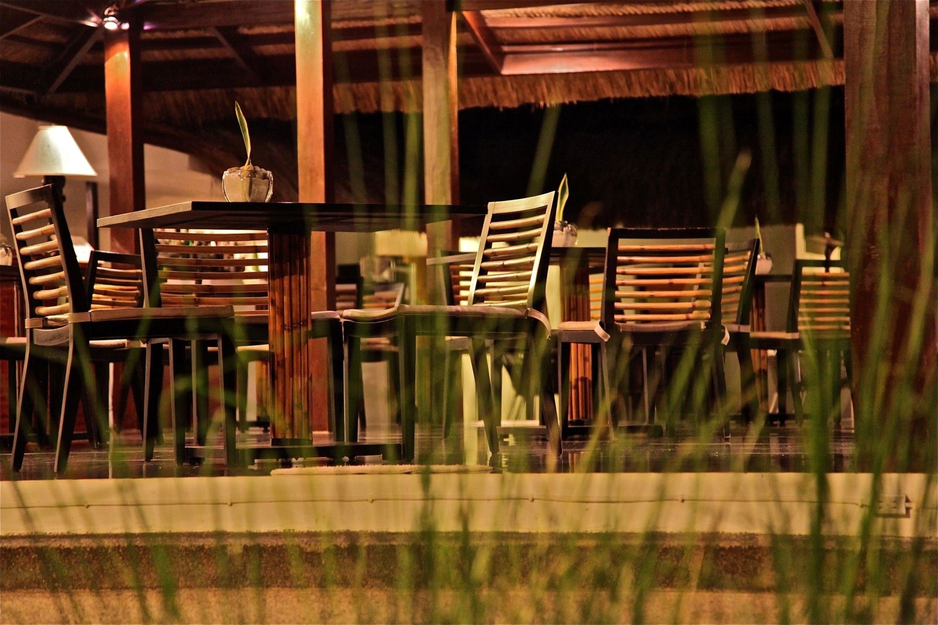 Een terras met houten tafels en stoelen, een rieten dak en een lamp.