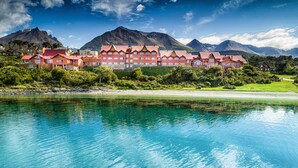 Een groot hotel met meerdere gebouwen met rode daken, gelegen aan een helderblauwe rivier met bergen op de achtergrond.
