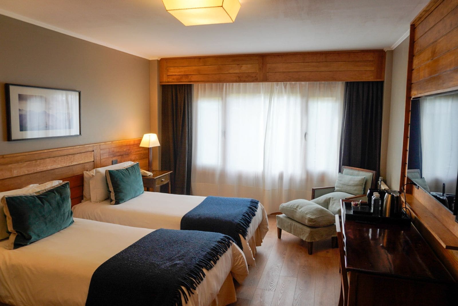 Habitación de hotel con dos camas, un escritorio, una silla, una lámpara y un ventanal con cortinas.