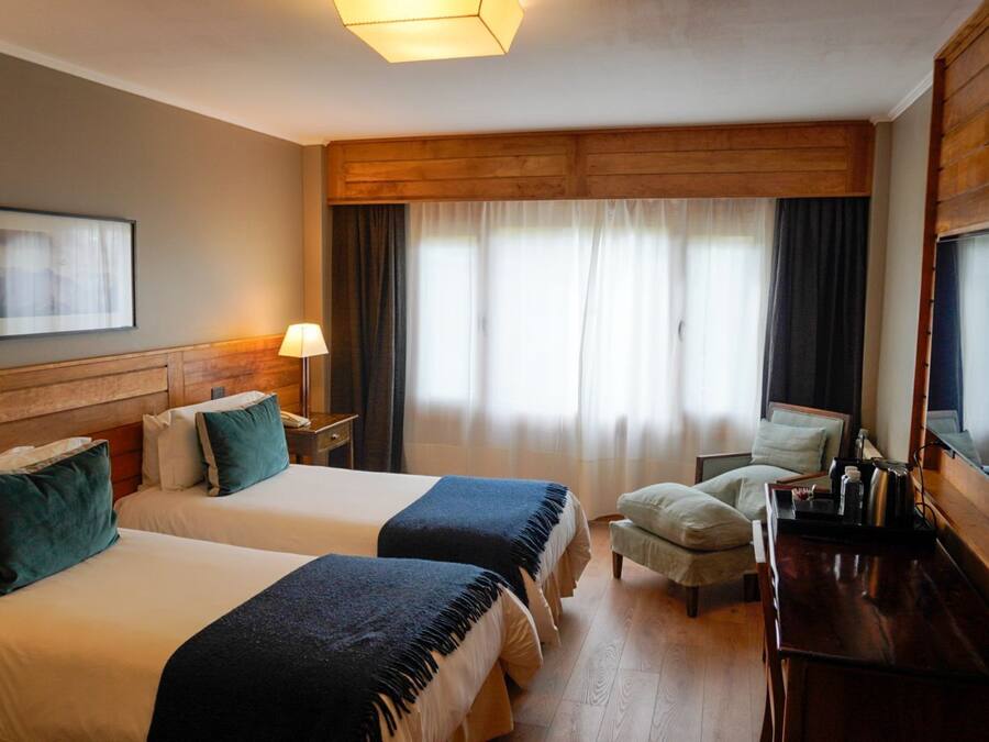Habitación de hotel con dos camas, un escritorio, una silla, una lámpara y un ventanal con cortinas.