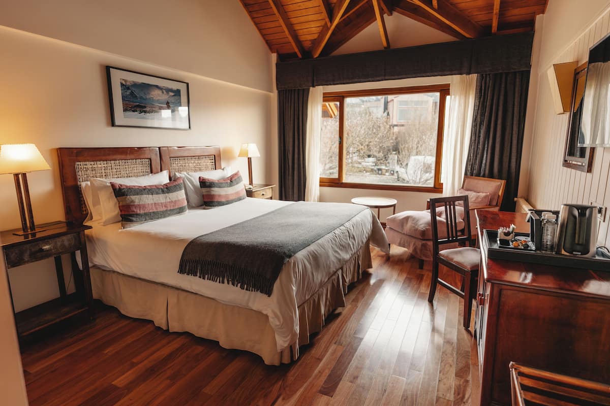 Un dormitorio con una cama grande, cabecera de madera, lámparas de noche, una mesita y una ventana con cortinas.