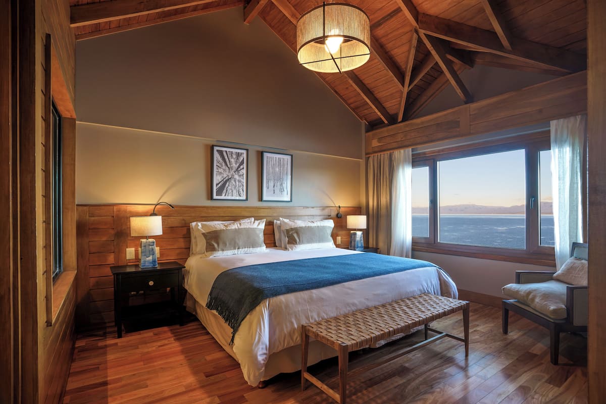 Un dormitorio con una cama grande, cabecera de madera, dos mesitas de noche con lámparas, un banco, una silla y un ventanal con vistas.