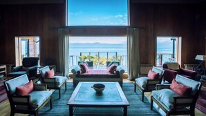 Amplia habitación con un ventanal que ofrece vistas al mar y a las montañas. Varias sofás y sillones están dispuestos alrededor de una mesa de centro.