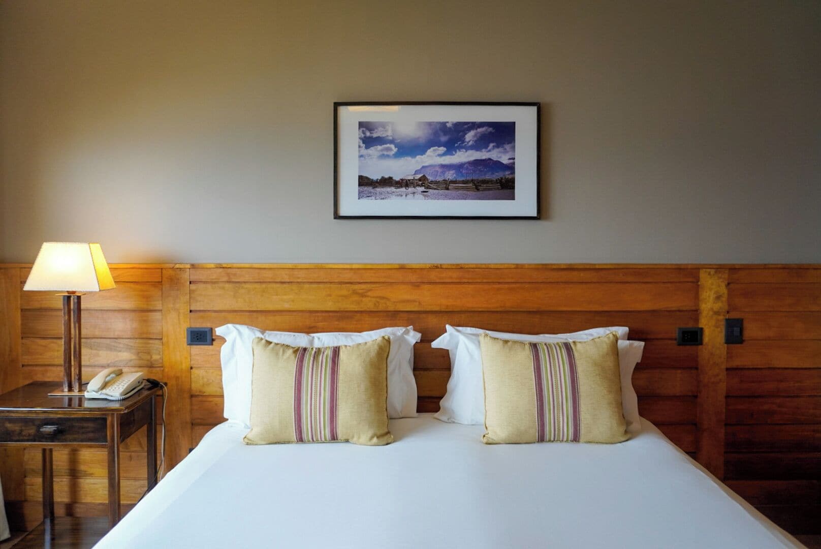 A hotel room with a wooden headboard, a bed with white linens, two striped pillows, a bedside table with a lamp and a phone, and a framed picture on the wall.