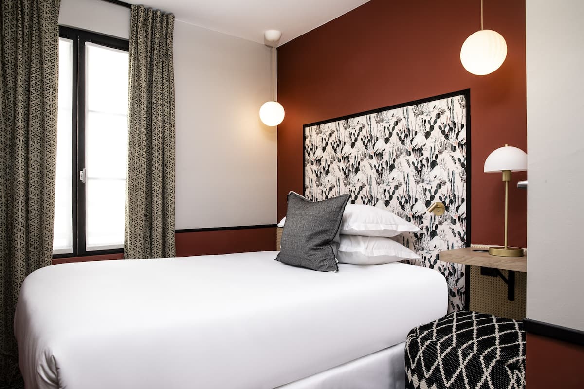 A hotel room with a large bed, a headboard with a floral pattern, a bedside table with a lamp, and a window with patterned curtains.
