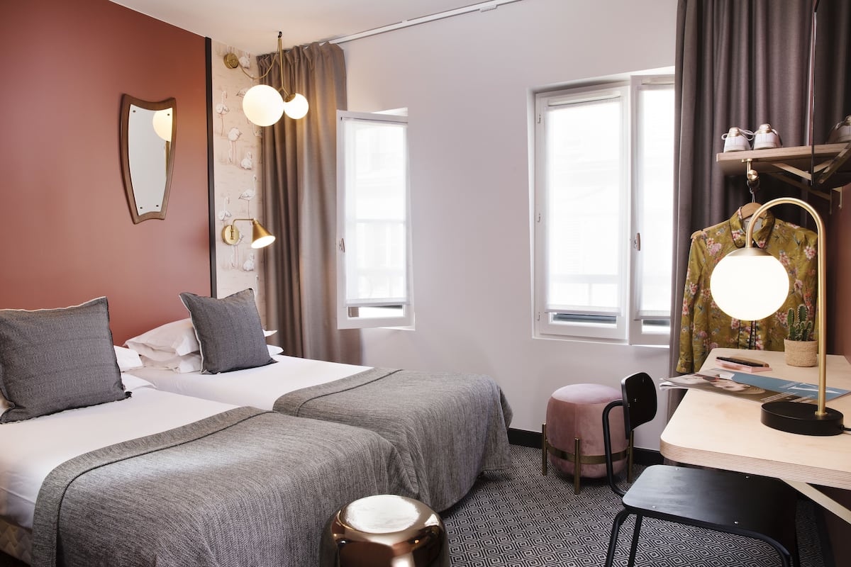 A hotel room with two beds, a desk with a chair, a vanity with a mirror, and a window with curtains.