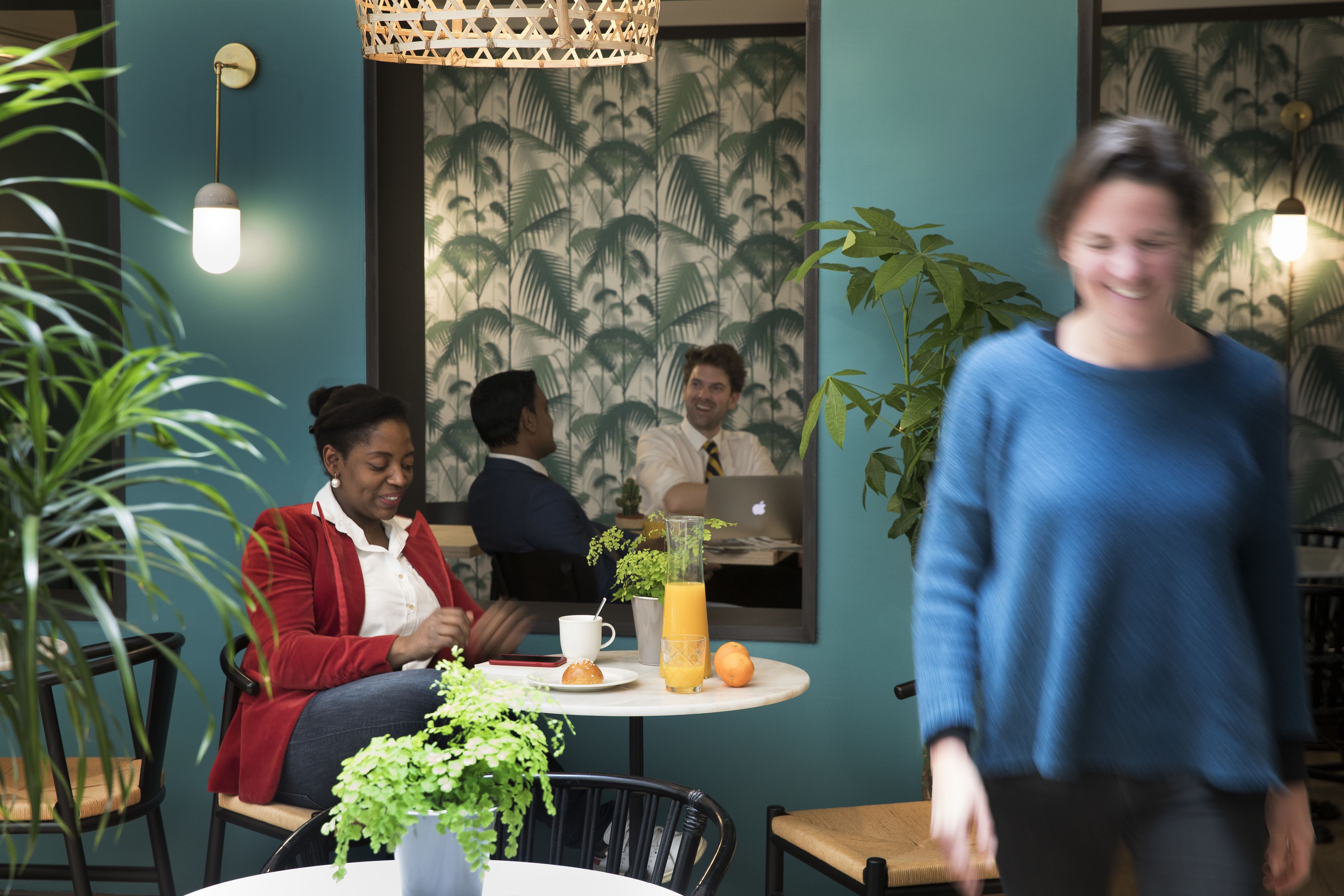 Un café avec un papier peint aux motifs tropicaux, une table ronde avec une boisson, et un luminaire suspendu.