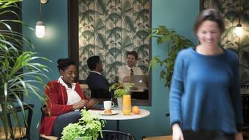 Un café avec un papier peint aux motifs tropicaux, une table ronde avec une boisson, et un luminaire suspendu.