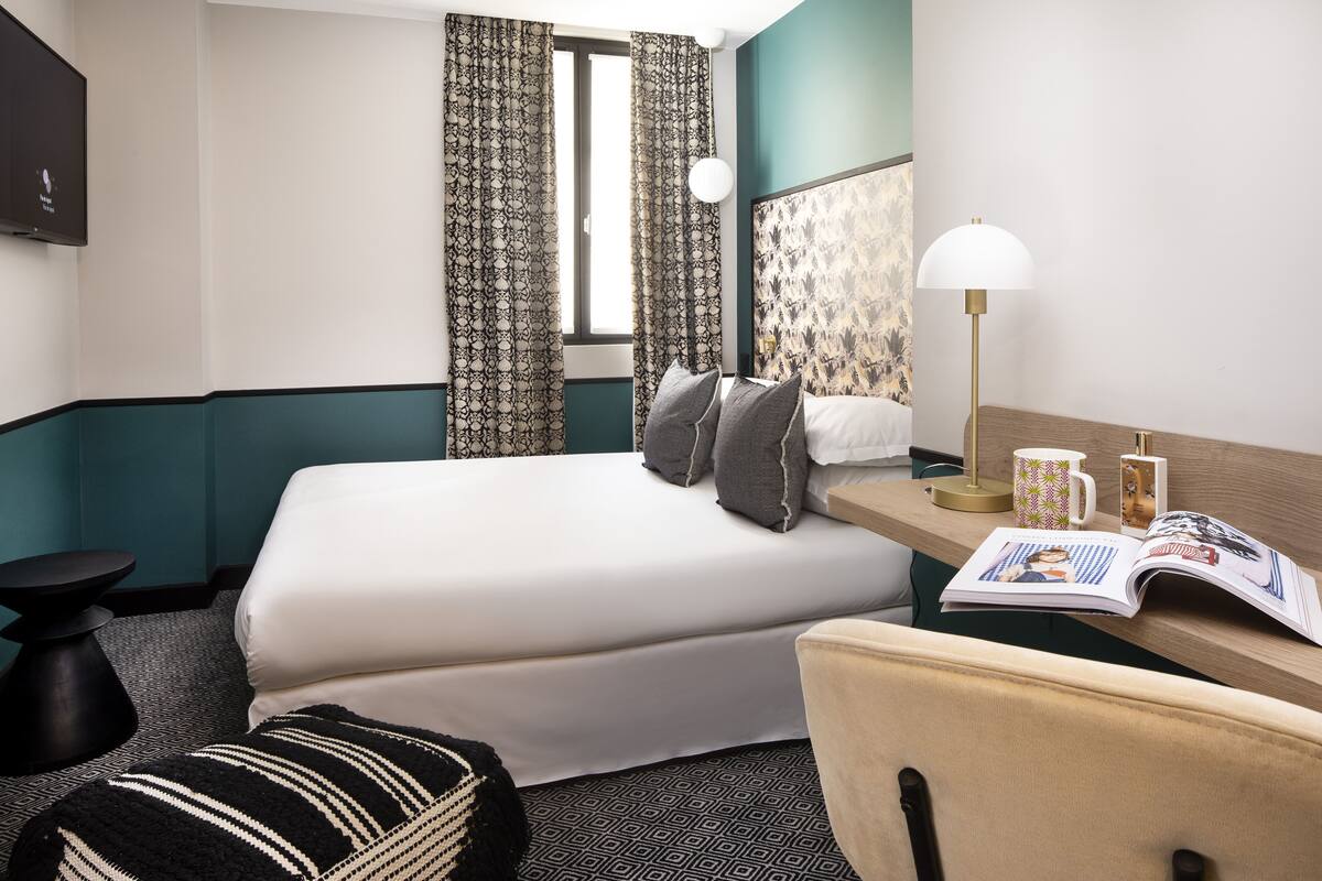 A hotel room with a bed, a desk with a lamp, a magazine, and a window with curtains.