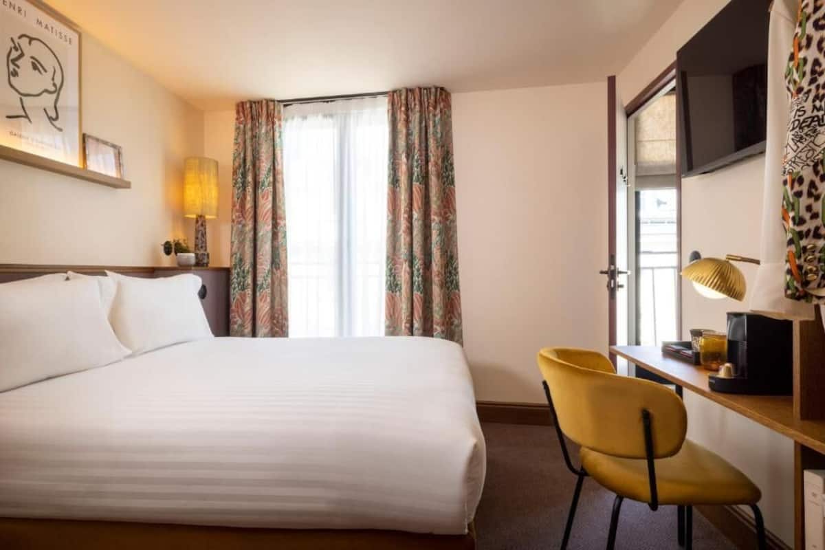 A hotel room with a bed, a desk with a chair, a lamp, and a window with curtains.