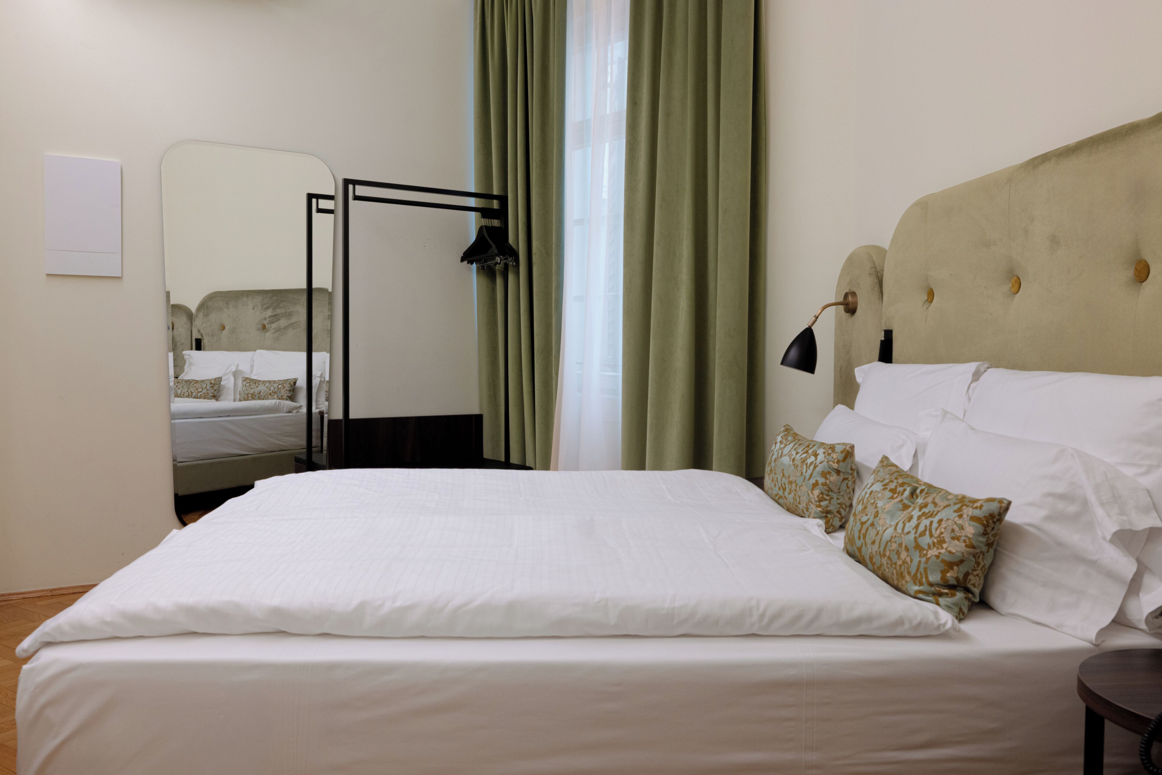 A neatly made bed with white linens, a headboard with buttons, a mirror, and a window with green curtains.