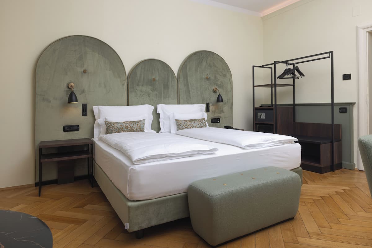 A double bed with a green upholstered bench, a small table, and a hanging clothes rack in a room with wooden flooring and green wall panels.