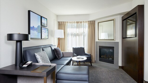 A modern hotel room with a grey sofa, a small armchair, a desk with a lamp, a fireplace, and framed pictures on the wall.