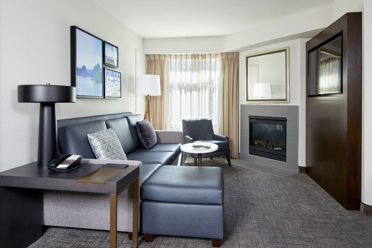 A modern hotel room with a grey sofa, a small armchair, a desk with a lamp, a fireplace, and framed pictures on the wall.