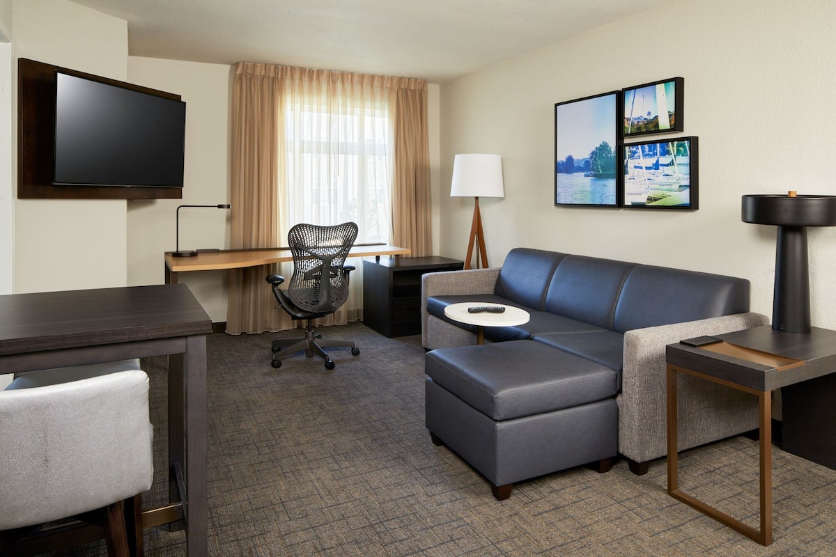 A hotel room with a flat-screen TV, a desk with a chair, a sofa, a lamp, and two framed pictures on the wall.