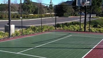 An outdoor basketball court with a green playing surface and a red surrounding area, surrounded by a fence and trees.