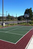 An outdoor basketball court with a green playing surface and a red surrounding area, surrounded by a fence and trees.