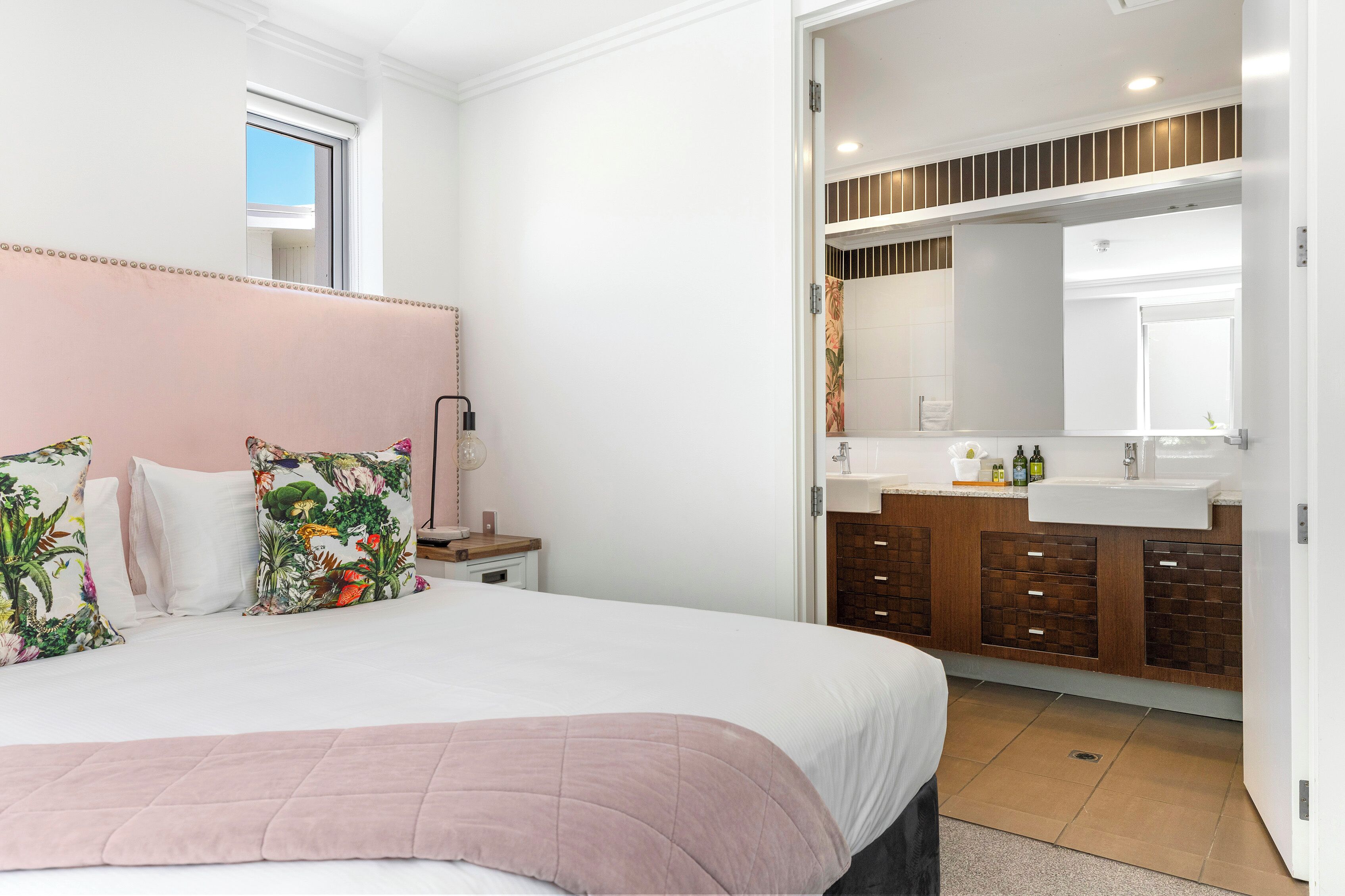 Une chambre à coucher avec un grand lit, une table de chevet, une commode et une salle de bain équipée d’un lavabo et d’un miroir.