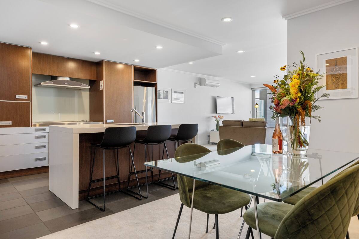 A modern kitchen with a glass dining table, bar stools, and a vase with flowers.