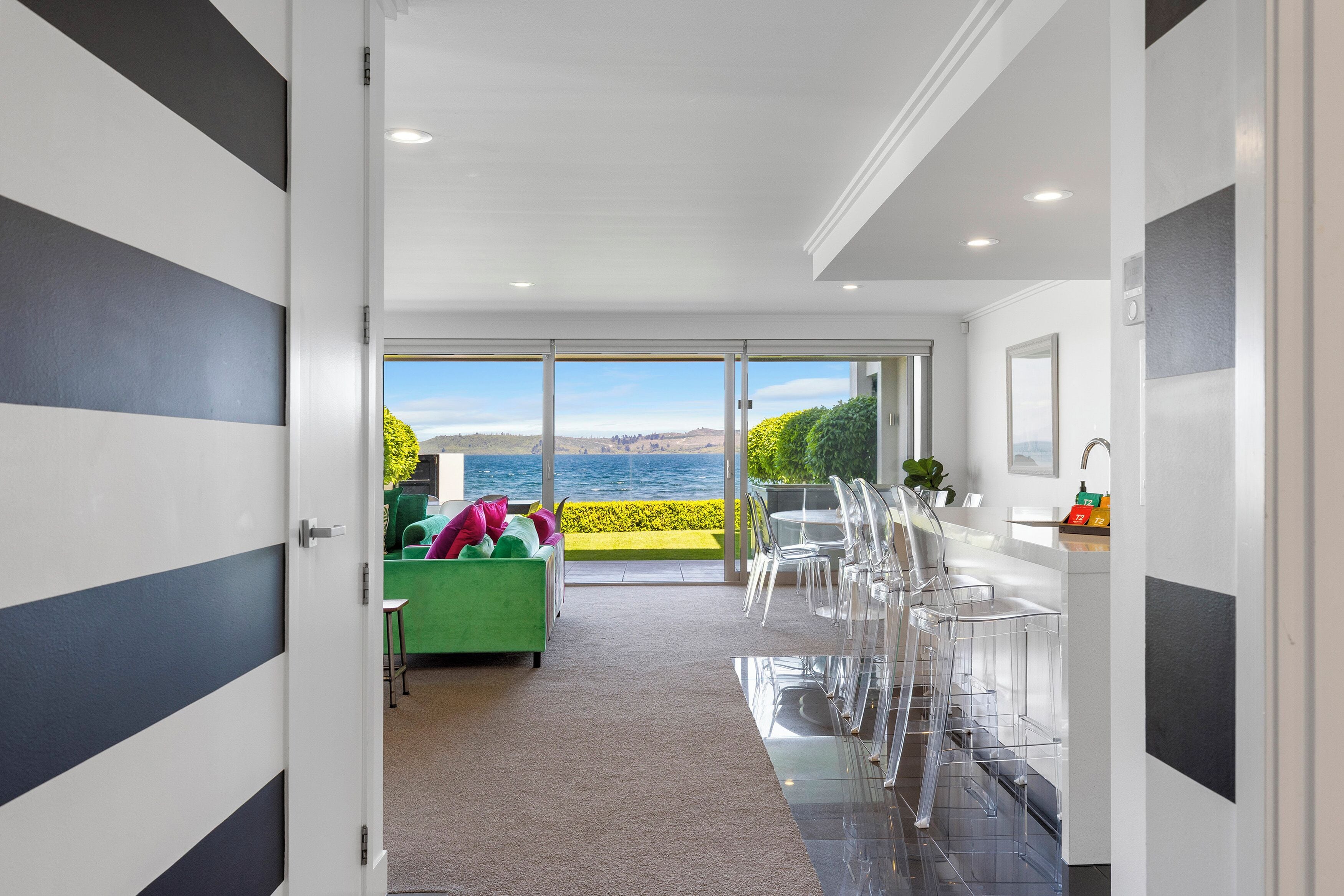 A modern interior with a view of the sea, a green sofa, clear chairs, and a white kitchen area.