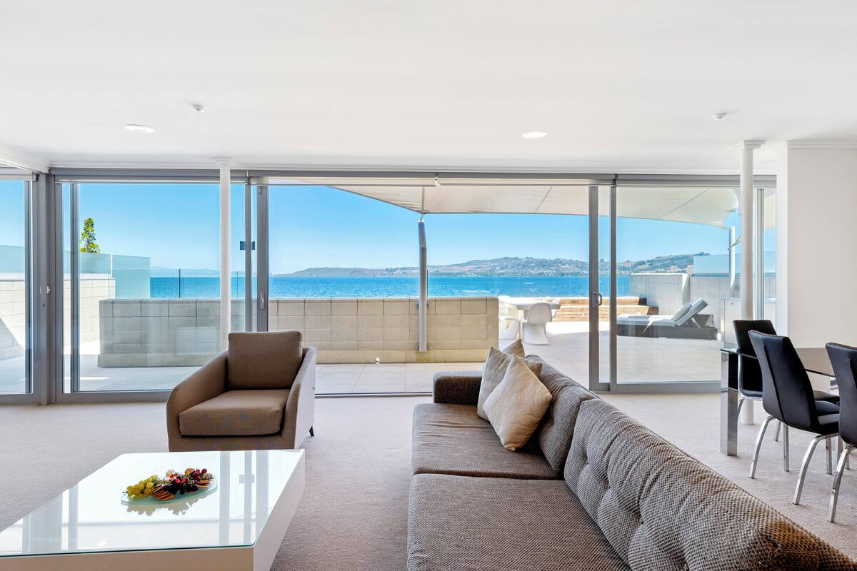 A modern living room with a glass wall, a sofa, a coffee table, and a view of the sea.