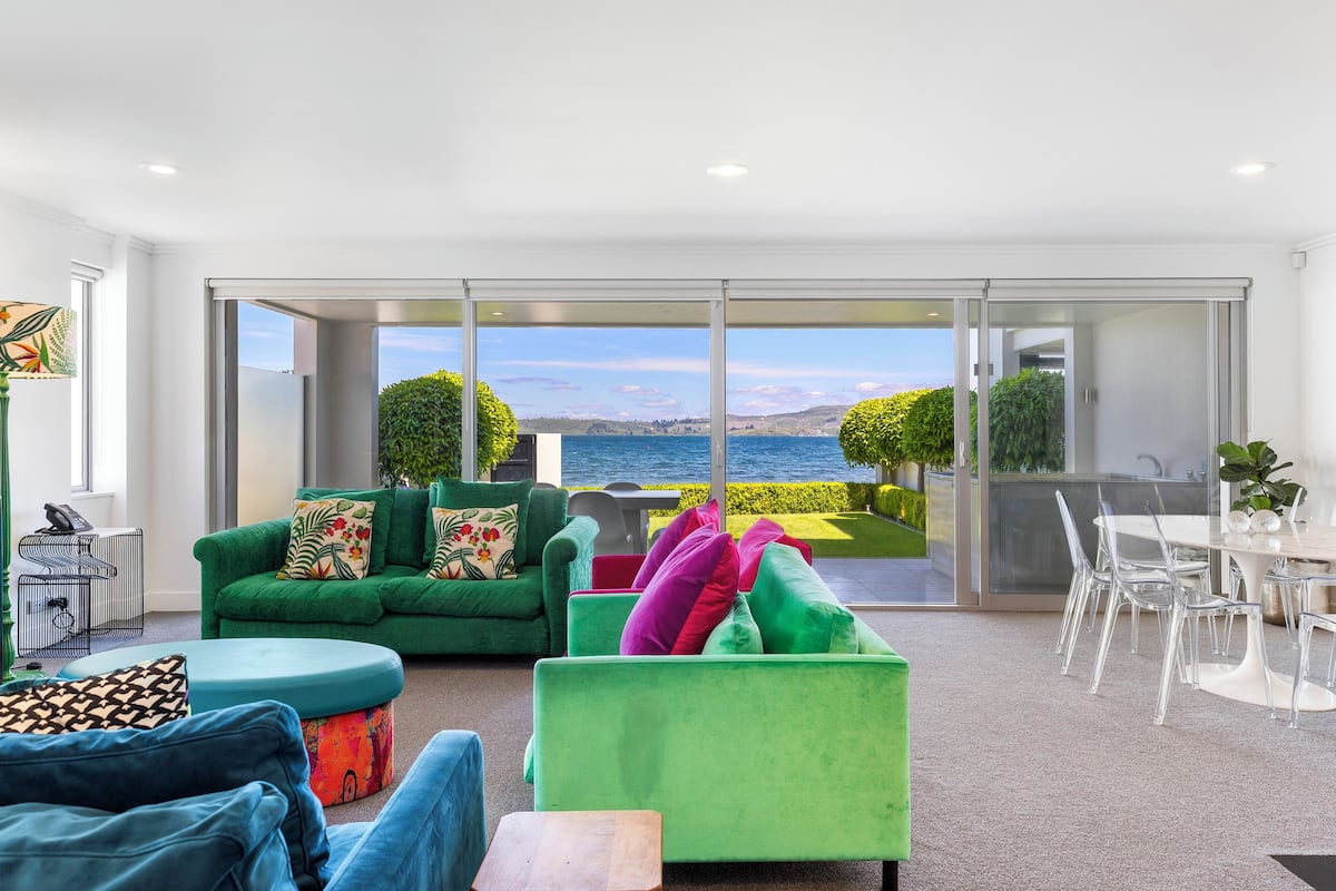 A modern living room with a view of the sea, green sofas, a dining area, and a large sliding glass door.