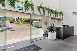 Zona de recepción con puerta de vidrio, mostrador de recepción, plantas de interior y vista despejada al exterior.