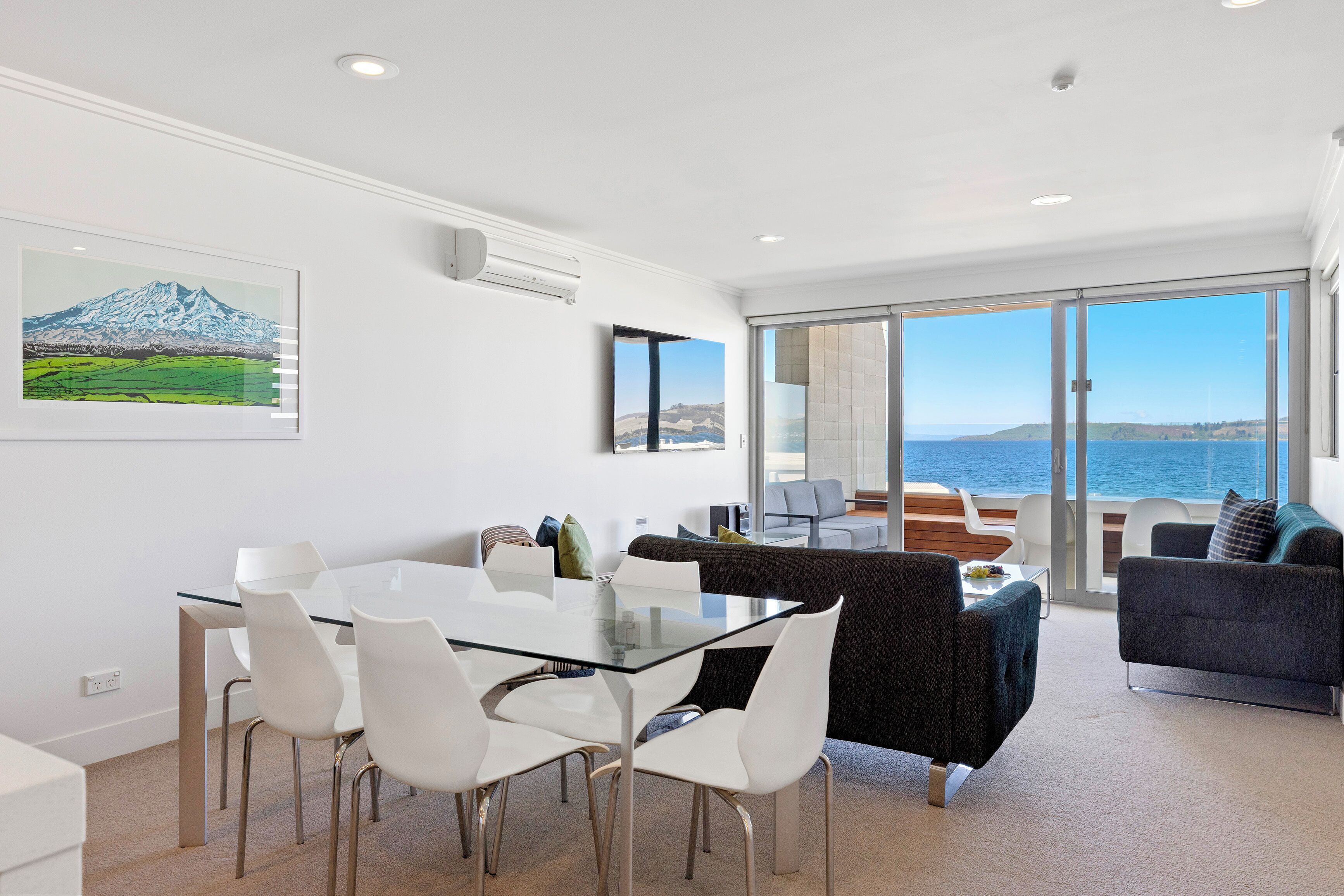 Un salon moderne avec une table en verre, des chaises blanches, un canapé foncé et une vue sur la mer.