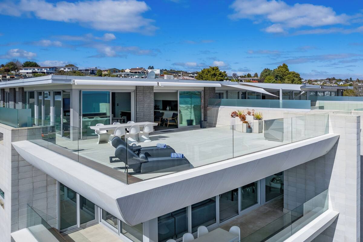A modern, multi-story residential building with large glass windows, a rooftop terrace with outdoor furniture, and a view of the surrounding neighborhood.