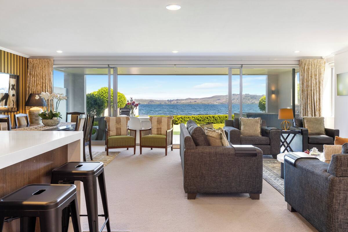 A modern living room with a large window overlooking a body of water, furnished with sofas and chairs, and a bar area with stools.