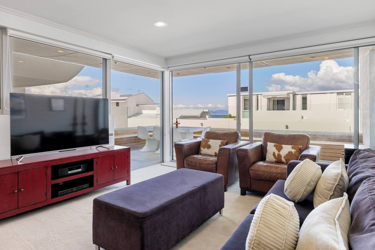A modern living room with a flat-screen TV, leather sofas, and a large window offering a view of the sky and neighboring buildings.