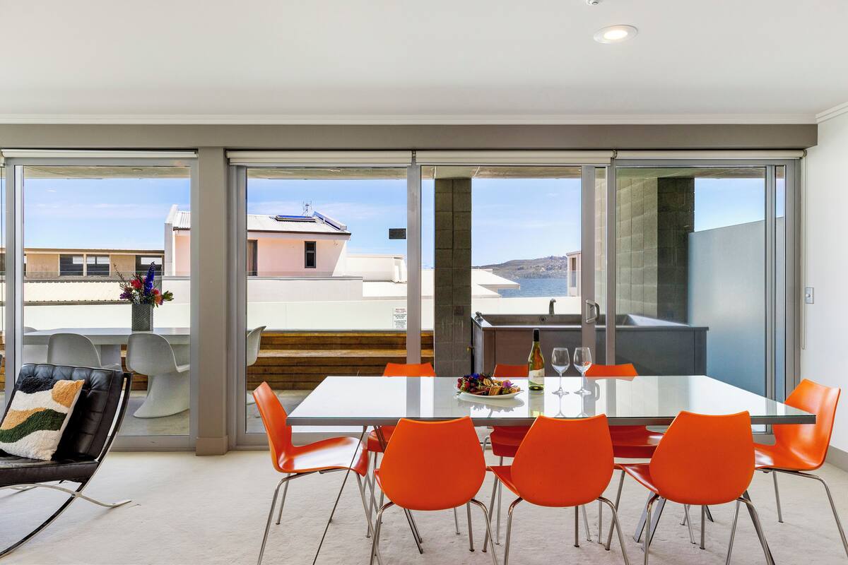 A modern dining area with a glass table, orange chairs, and a view of the sea through large windows.