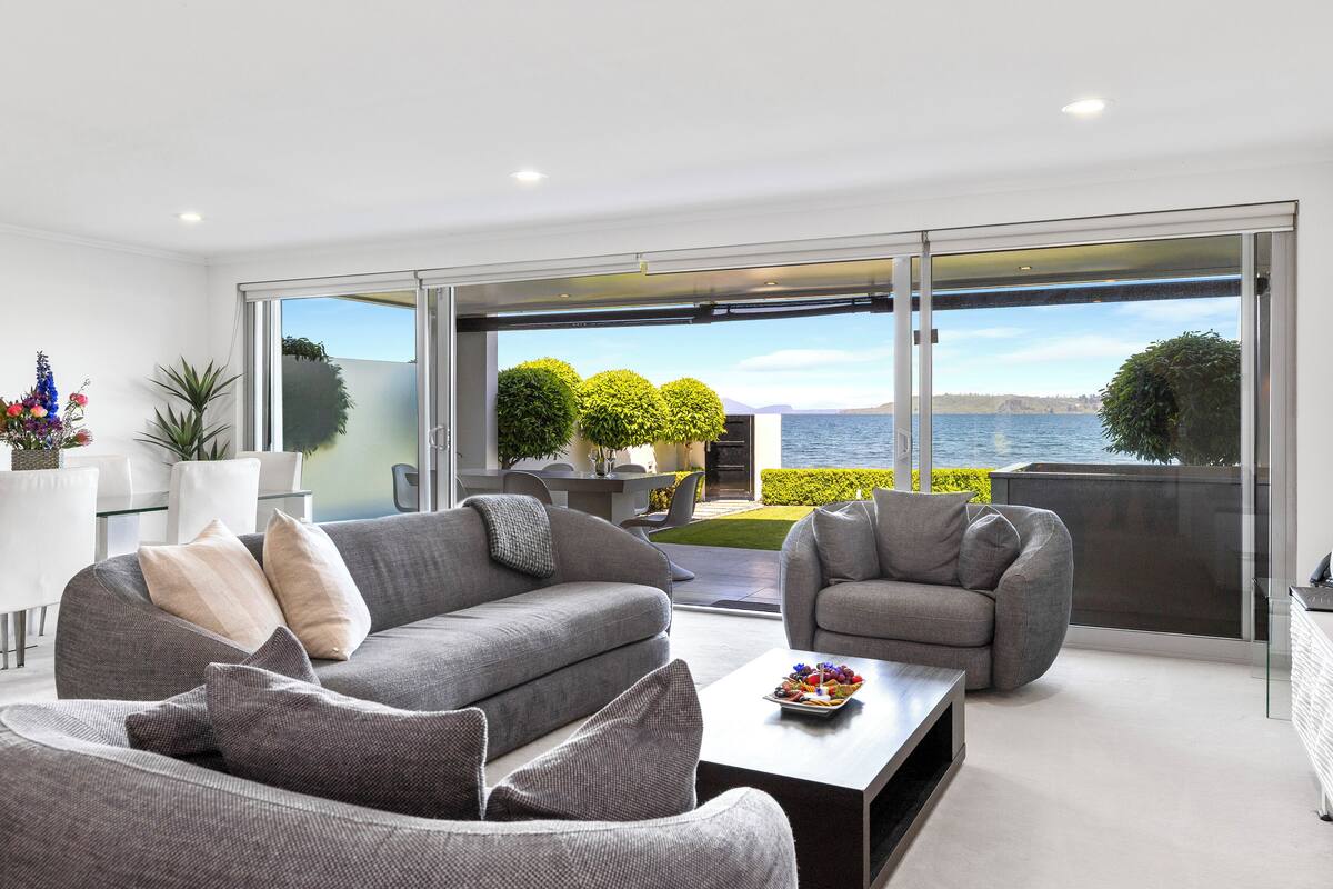A modern living room with a grey sofa, a coffee table, and a view of the sea through large sliding glass doors.