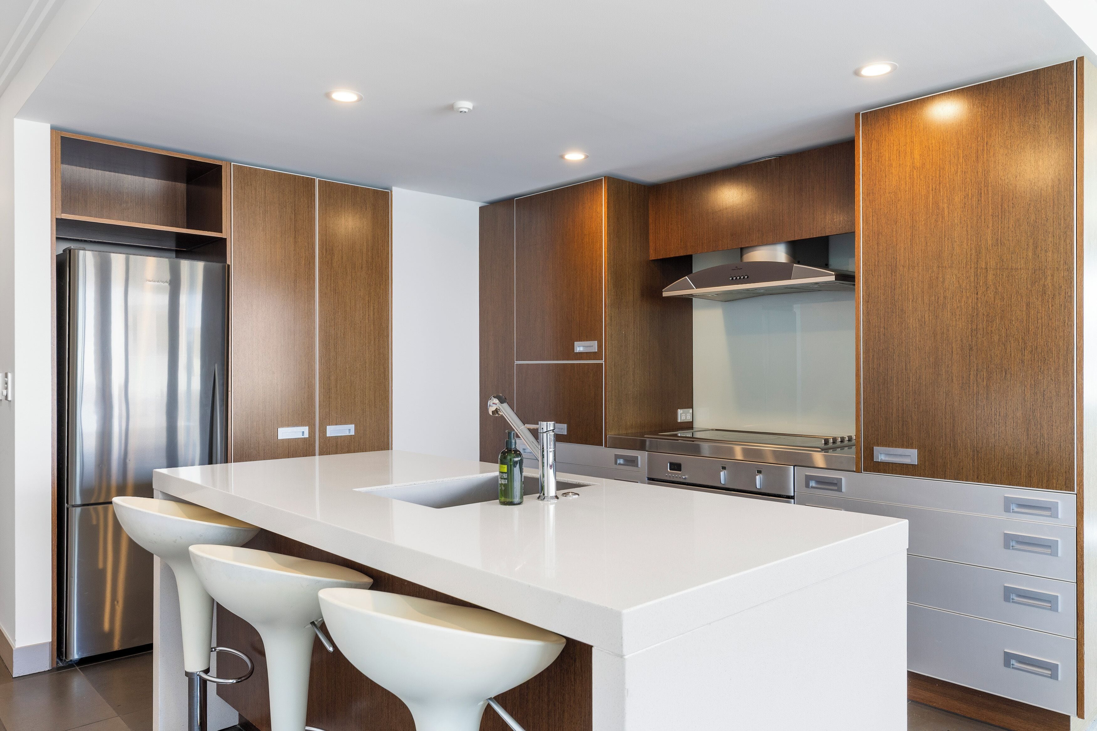 A modern kitchen with a stainless steel refrigerator, a large island with bar stools, wooden cabinetry, and a stainless steel range hood.