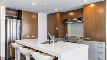 A modern kitchen with a stainless steel refrigerator, a large island with bar stools, wooden cabinetry, and a stainless steel range hood.