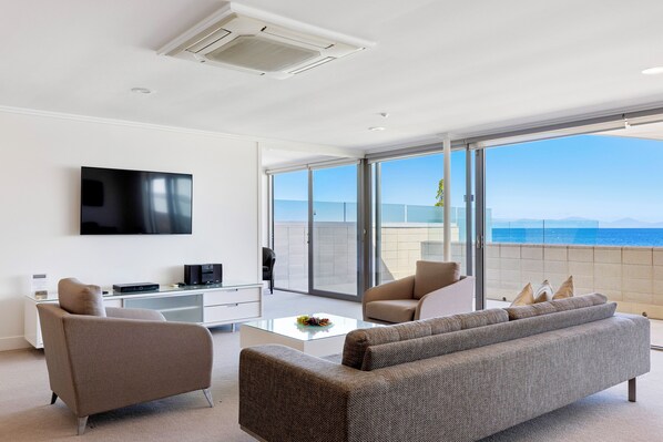 A modern living room with a flat-screen TV, a sofa, a coffee table, and a balcony with a view of the sea.