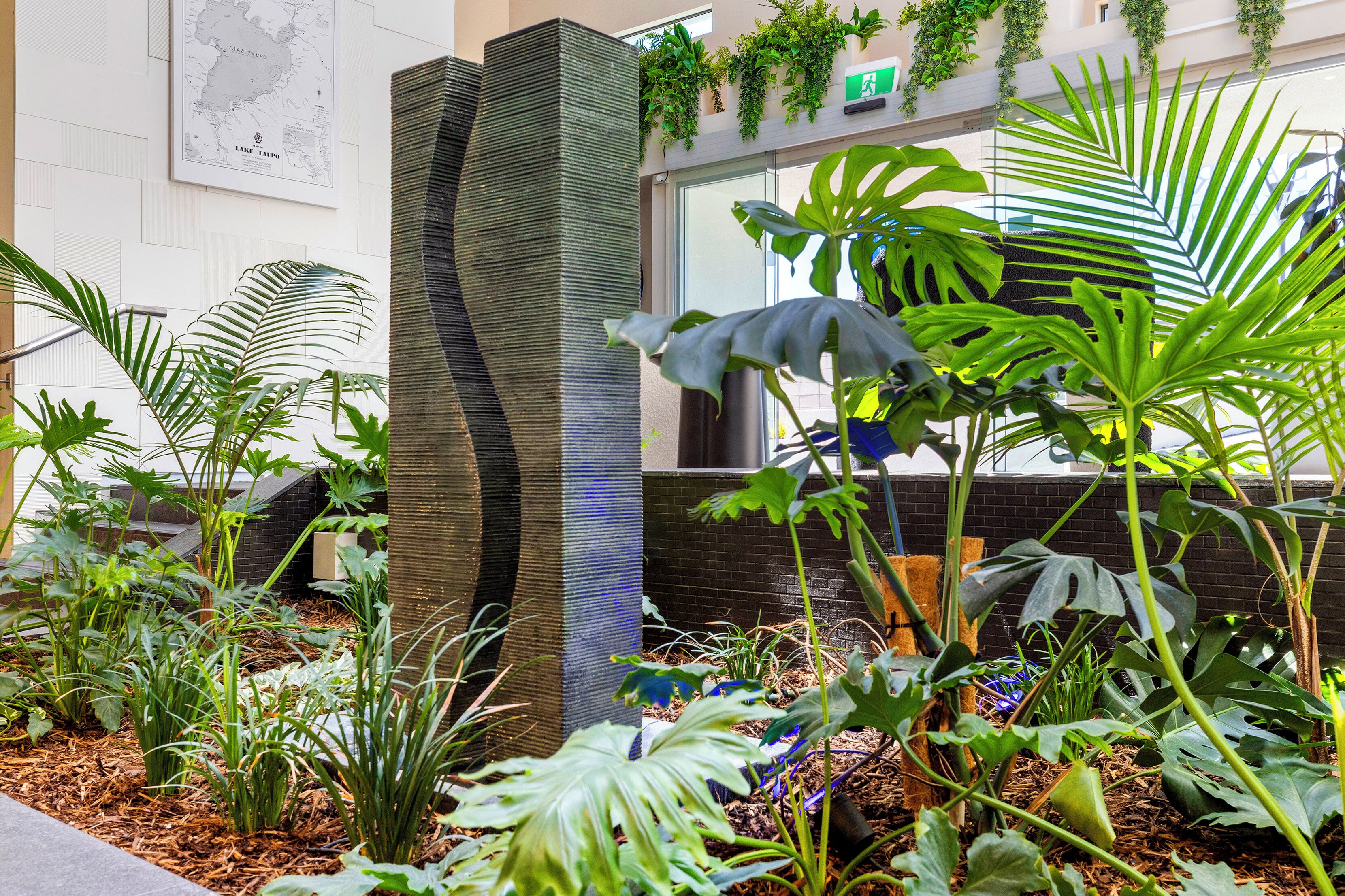 Interior de un edificio moderno con una columna alta y texturizada, plantas de interior y un mapa en la pared.