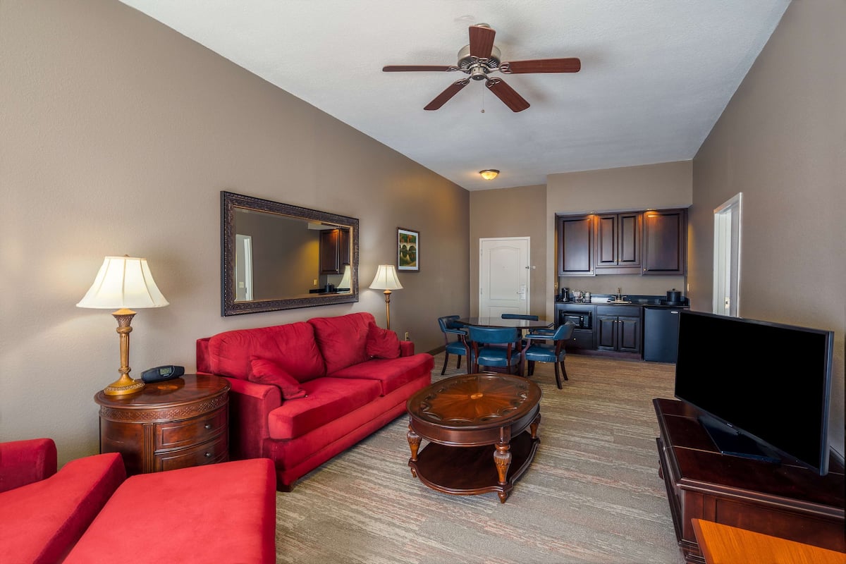 A living room with a red sofa, a coffee table, a TV, and a ceiling fan. There is a kitchen in the background.