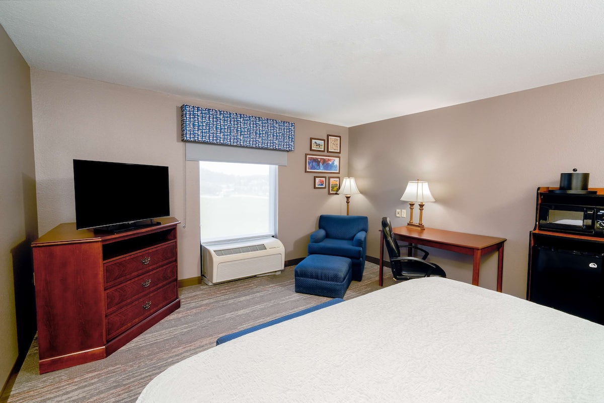 A hotel room with a bed, a TV on a wooden dresser, a desk with a lamp, a chair, and a small refrigerator.