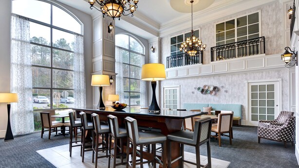 A spacious dining area with a large island, multiple chairs, and a view of the outdoors through large windows. - Hampton Suites Birmingham Hoover (Birmingham)