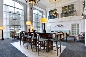 A spacious dining area with a large island, multiple chairs, and a view of the outdoors through large windows. - Hampton Suites Birmingham Hoover (Birmingham)