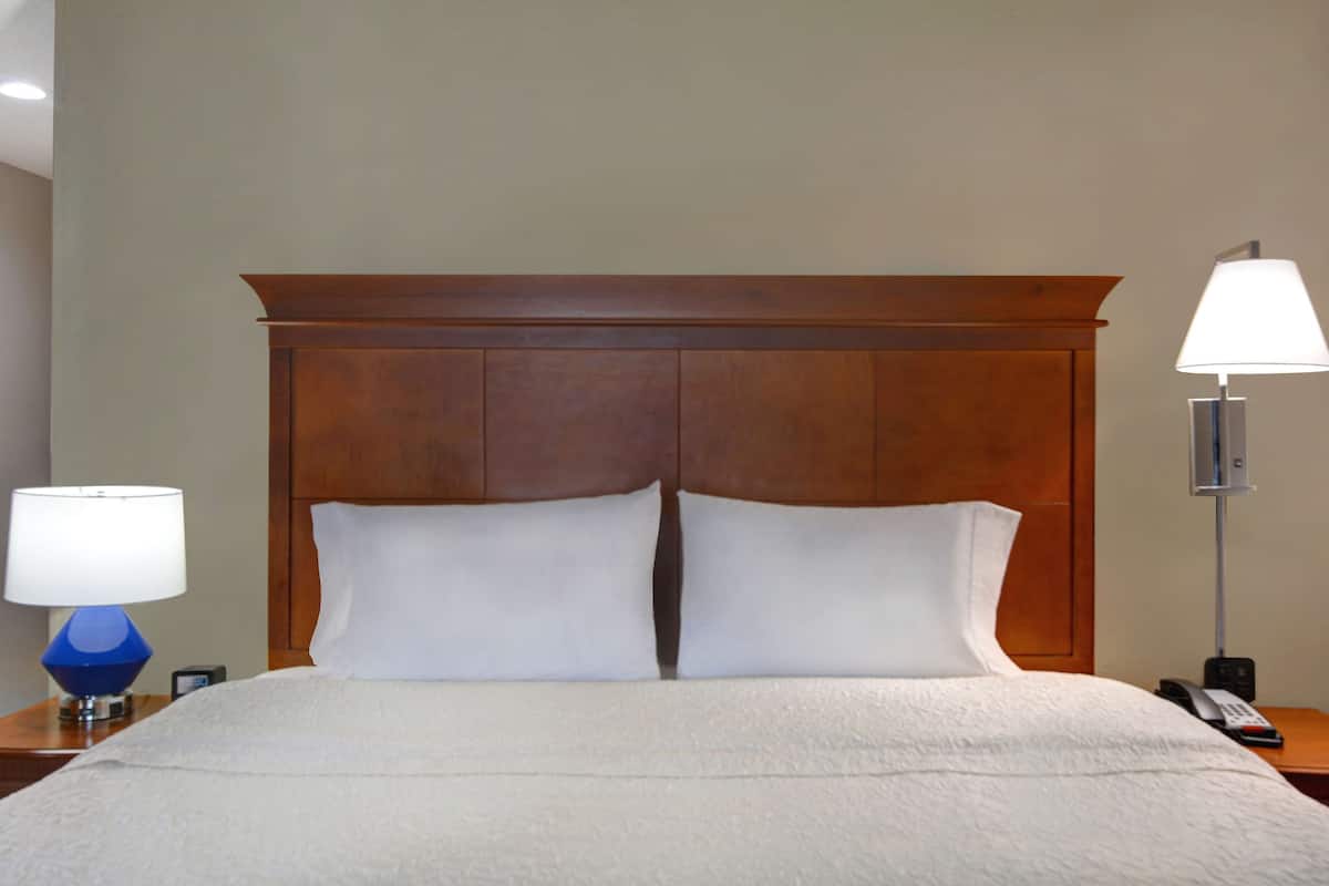 A hotel room with a wooden headboard, a white bedspread, two white pillows, a blue lamp, and a white lamp on a nightstand.