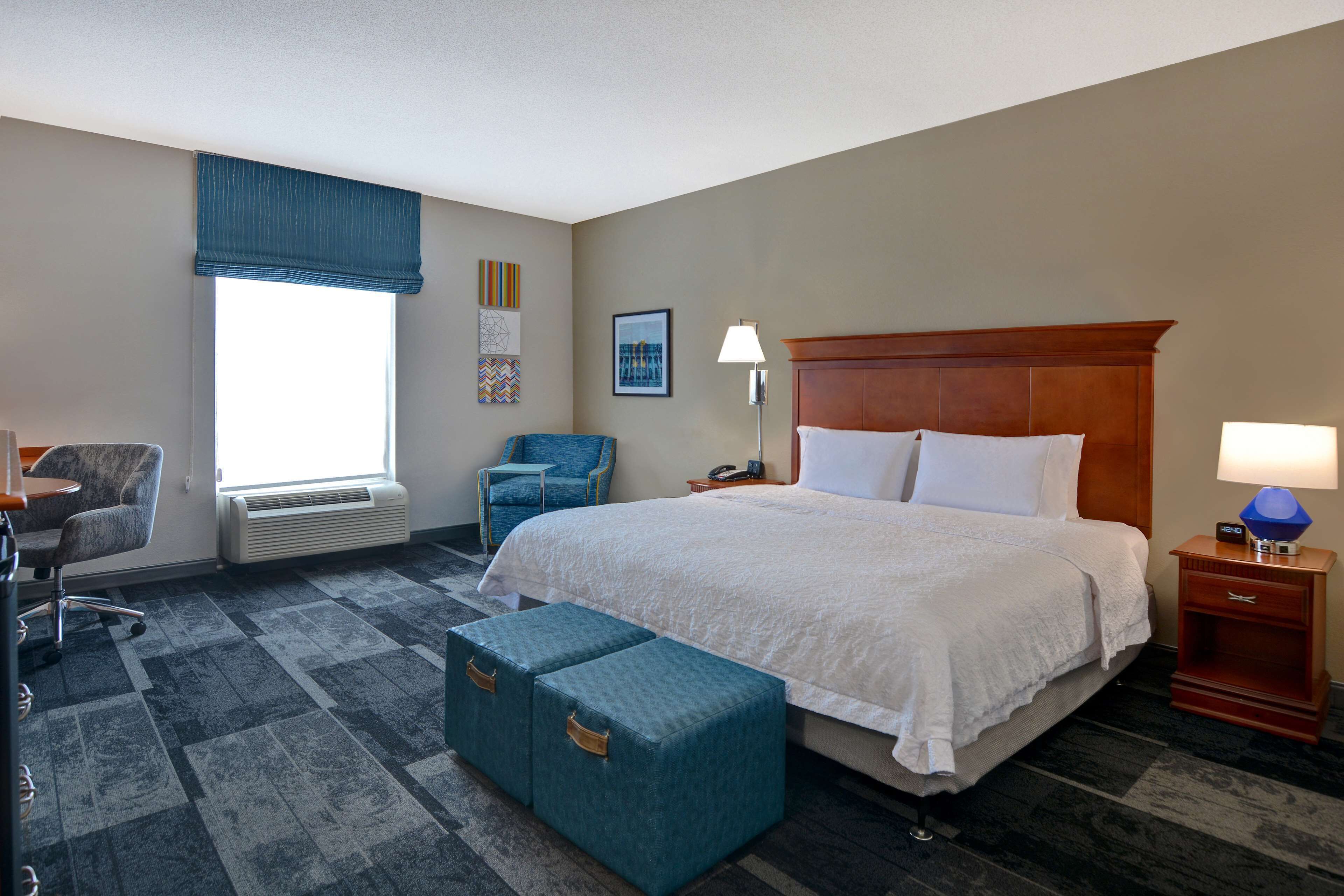 A hotel room with a large bed, a desk with a chair, a blue armchair, a bedside table with a lamp, and a window with blinds.