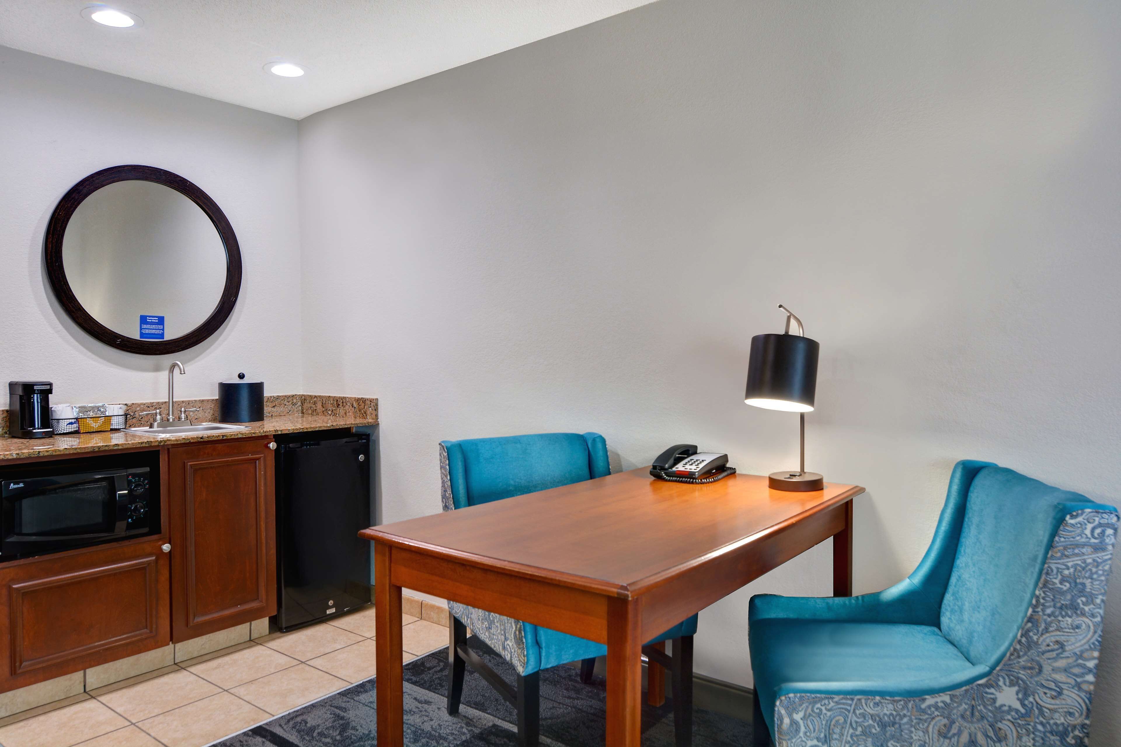 A hotel room with a wooden table, two blue upholstered chairs, a round mirror, a coffee maker, a phone, and a lamp.