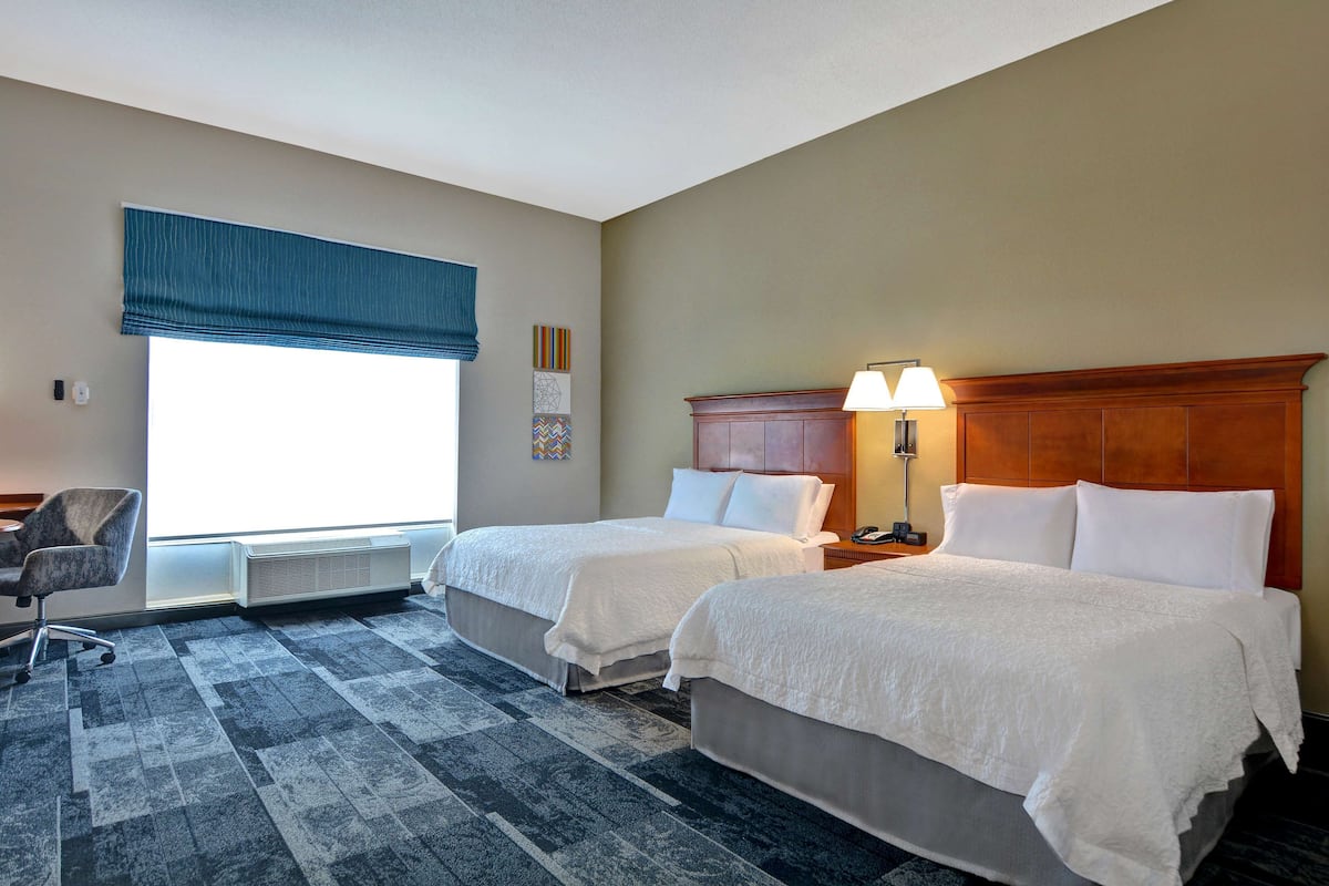 A hotel room with two beds, a desk, a chair, a lamp, and a window with blinds.