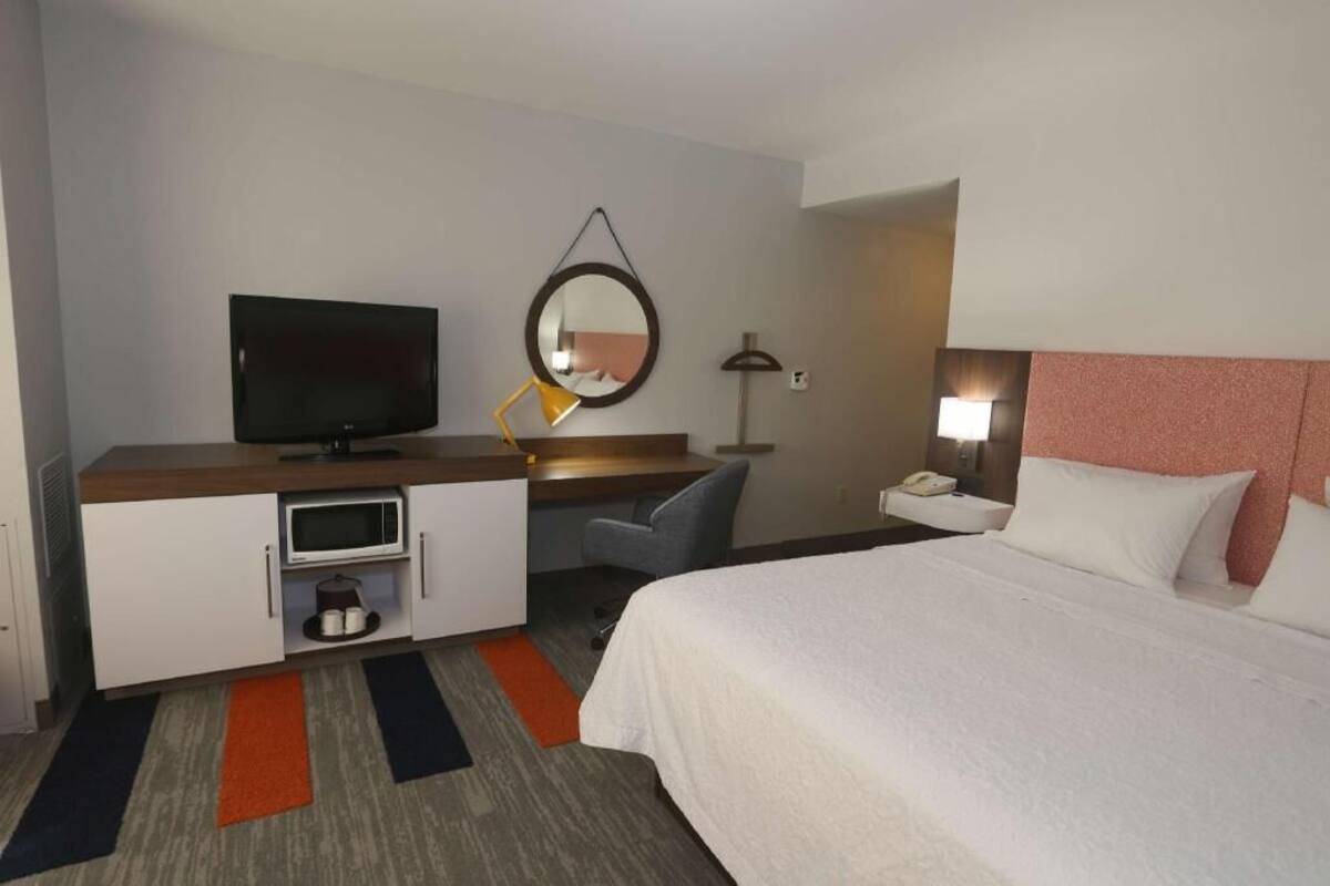 A hotel room with a bed, a desk with a TV, a microwave, a chair, a nightstand, and a mirror.
