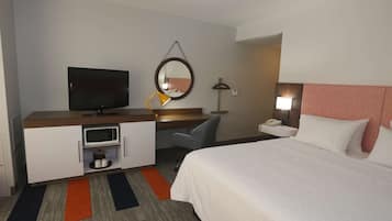 A hotel room with a bed, a desk with a TV, a microwave, a chair, a nightstand with a lamp, and a mirror on the wall.